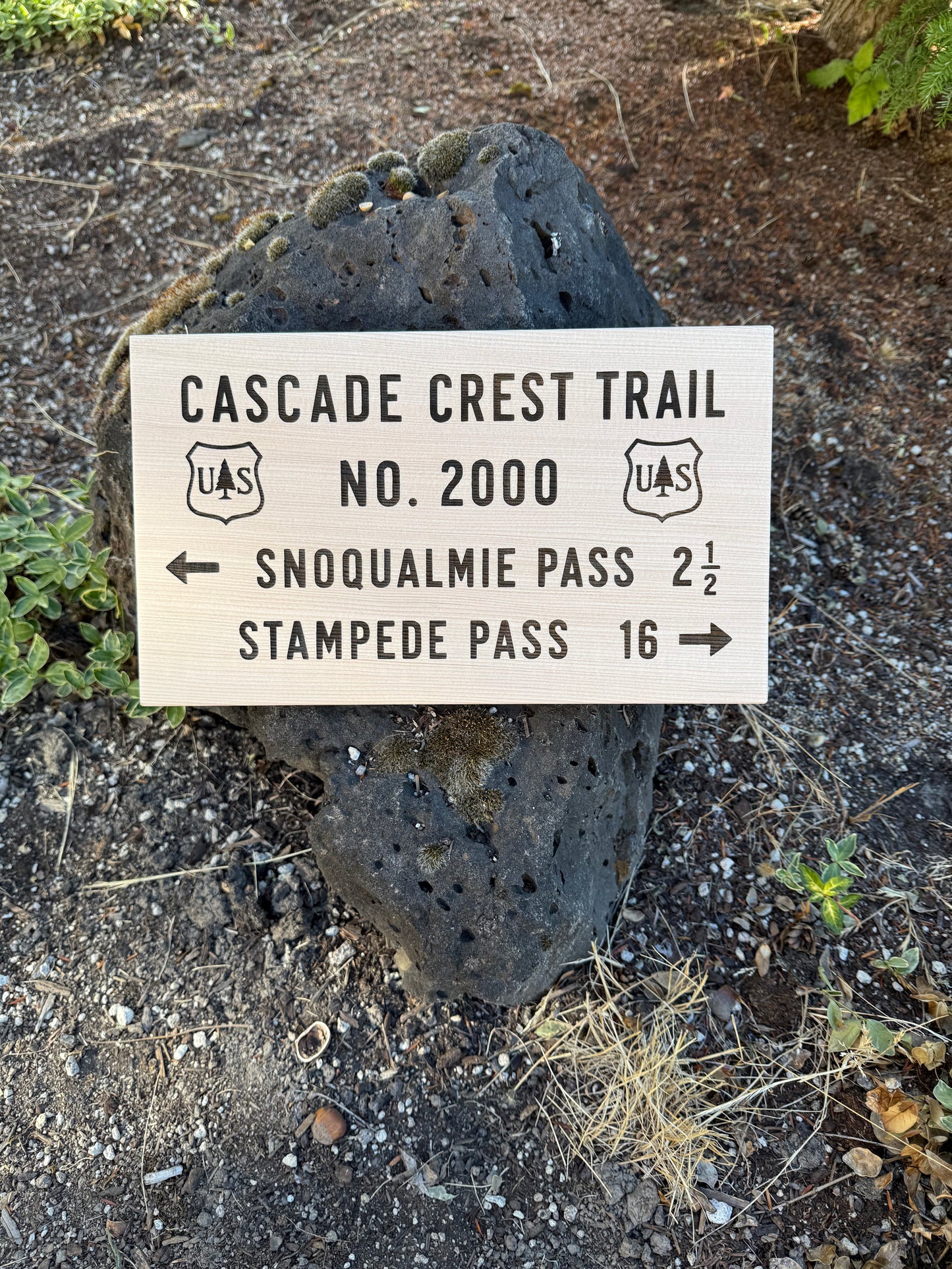 A rectangular wooden trail sign reading 'CASCADE CREST TRAIL No. 2000' with directions to Snoqualmie Pass and Stampede Pass, placed in front of a large dark volcanic rock on a dirt path outdoors.