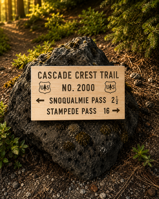 A rectangular wooden trail sign for Cascade Crest Trail, No. 2000, mounted on a large dark rock in a forest setting.