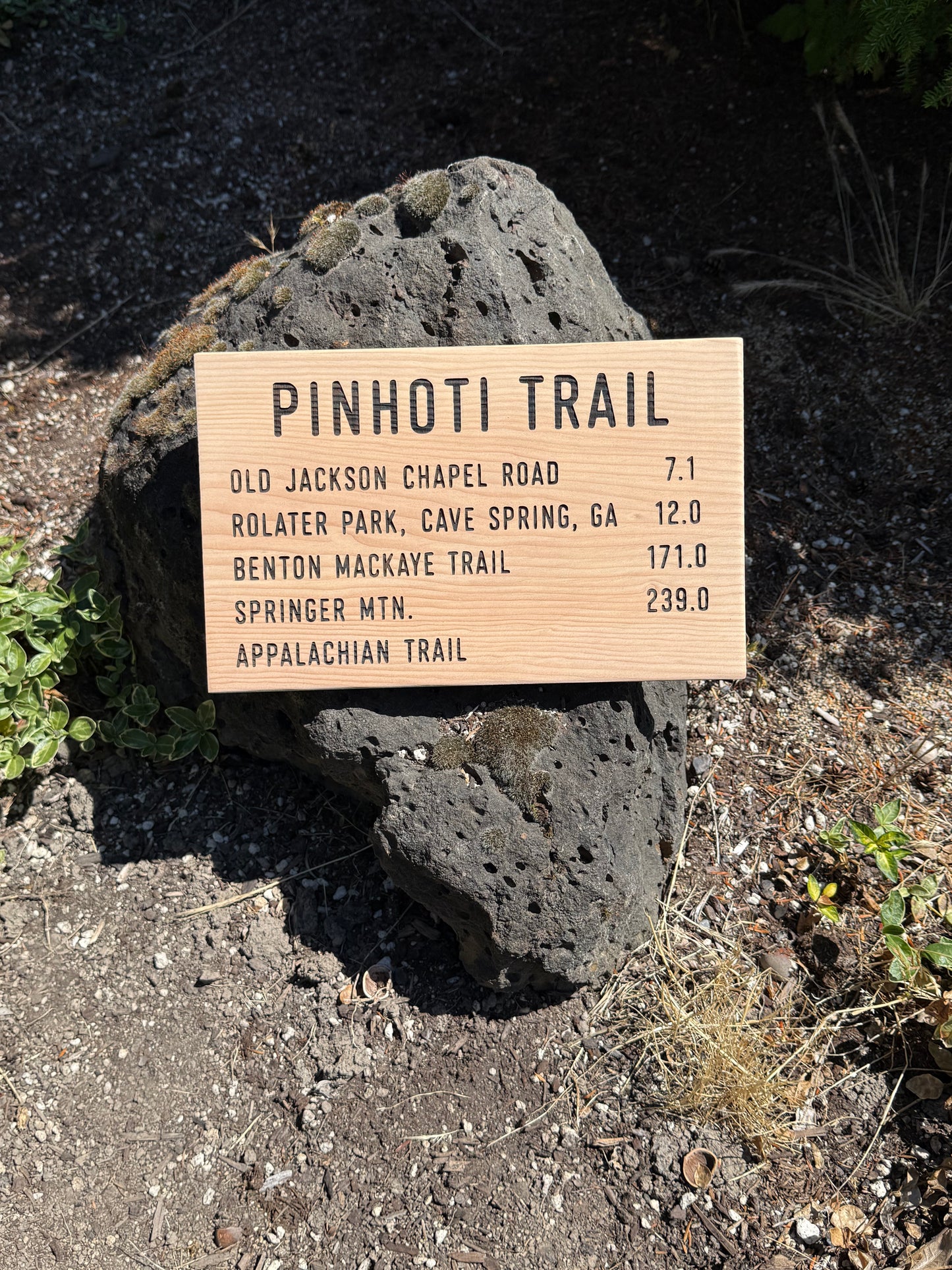 Wooden trail sign on a rock with trail distances listed, set against a natural outdoor background.