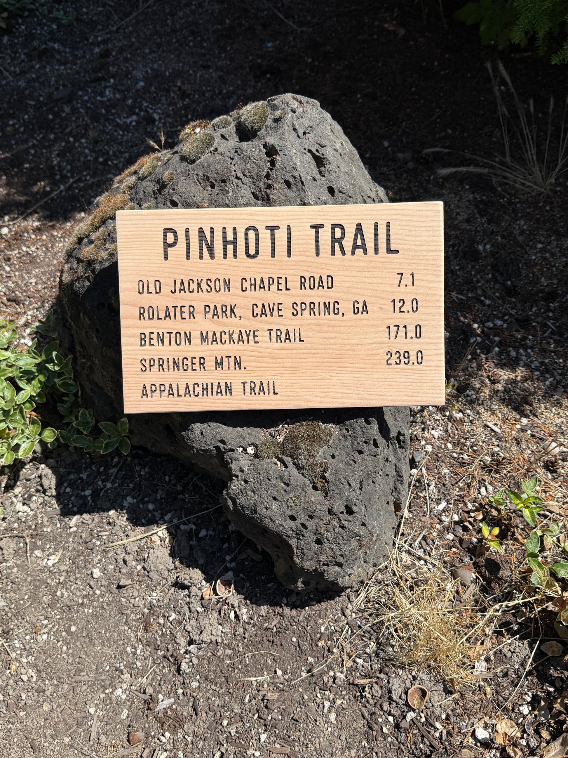 Wooden trail sign on a rock with trail distances listed, set against a natural outdoor background.