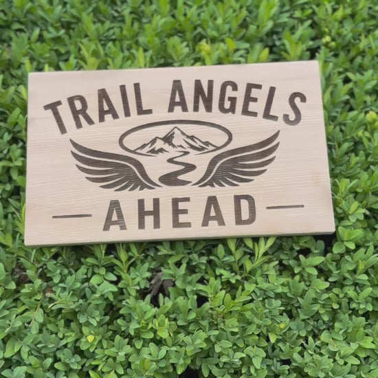 Trail Angels Ahead wood sign on a forest tree with hikers enjoying breakfast and trail magic in the background