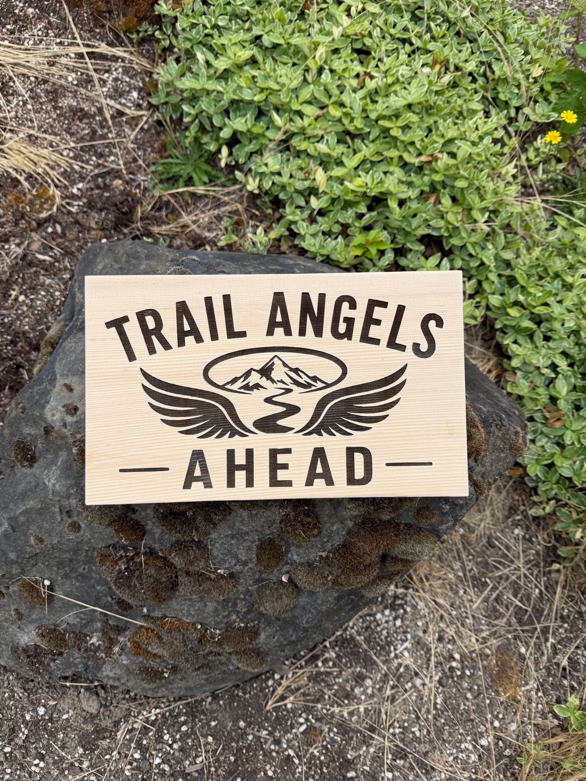 Rustic Trail Angel wooden sign displayed beside a hiking trail leading to a welcoming trail magic campsite.